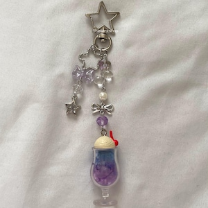 May include: A silver keychain with a star-shaped top, adorned with purple beads, bows, and a miniature sundae charm. The sundae features a blue and purple base, topped with vanilla ice cream and a red cherry.
