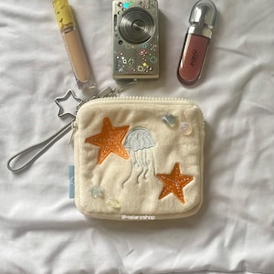 May include: An assortment of beauty and accessory items on a white surface. Includes a beige cosmetic tube, a silver digital camera, a Kiko lip gloss, and a cream-colored pouch with orange star and jellyfish embroidery. The pouch has a star-shaped zipper pull.