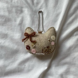 May include: A beige cat-shaped keychain made of felt, adorned with a brown bow, various buttons, and small star-shaped embellishments. The keychain has a silver chain and a small metal tag. The text "@4starzshop" is visible.