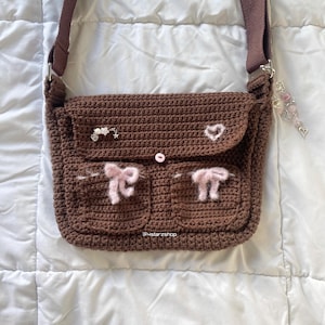 May include: A brown crochet shoulder bag with a flap and two front pockets. The bag features pink bow accents, a heart, and a flower embellishment. It has a brown strap and a decorative charm. The text "@ustarzshop" is visible.