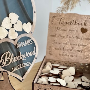 May include: Wooden guest book box with a heart-shaped frame and a sign that reads "Guestbook Please sign a and drop it into the frame So we will never forget our loved ones who came". The box is filled with wooden hearts for guests to sign.