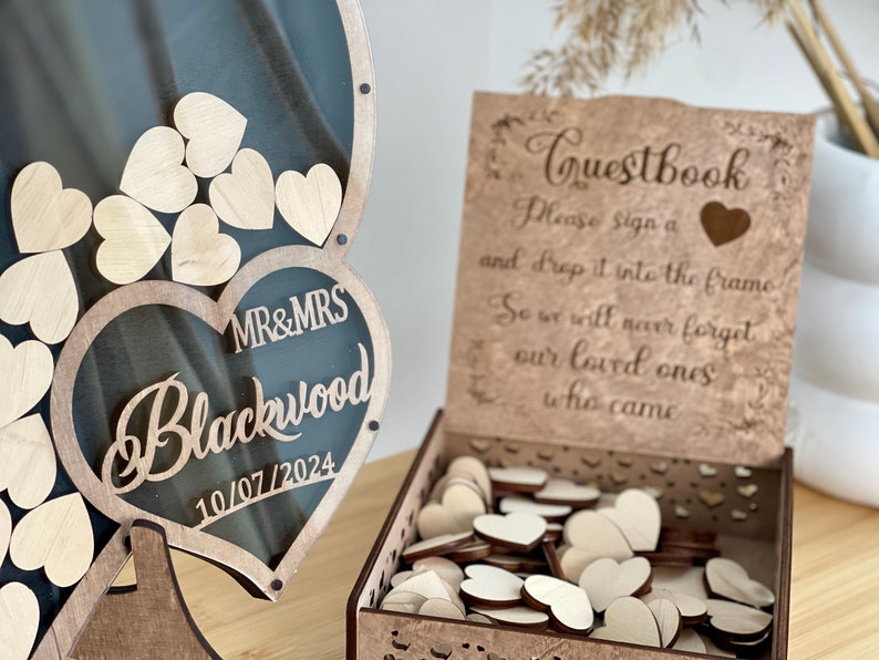 May include: Wooden guest book box with a heart-shaped frame and a sign that reads "Guestbook Please sign a and drop it into the frame So we will never forget our loved ones who came". The box is filled with wooden hearts for guests to sign.