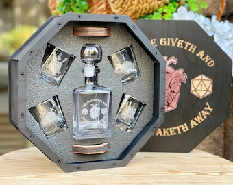 May include: A black wooden decanter set with a clear glass decanter and four matching glasses. The decanter and glasses are etched with a design. The set is displayed in a black octagonal box with the text "THE DICE GIVETH AND TAKETH AWAY".