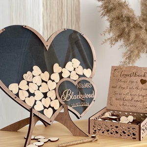 May include: Wooden heart-shaped guest book frame with a clear acrylic panel. The frame is on a stand and has a small heart-shaped sign with the text "MR&MRS Blackwood 10/07/2024". A separate wooden box with a heart-shaped cutout design contains additional wooden hearts for guests to sign.
