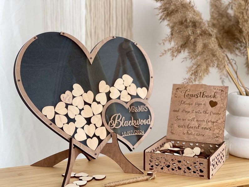 May include: Wooden heart-shaped guest book frame with a clear acrylic panel. The frame is on a stand and has a small heart-shaped sign with the text "MR&MRS Blackwood 10/07/2024". A separate wooden box with a heart-shaped cutout design contains additional wooden hearts for guests to sign.
