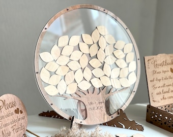 Puede incluir: Un árbol de madera para libro de visitas con un círculo transparente de acrílico alrededor. El árbol tiene hojas que los invitados pueden firmar y dejar caer en la caja. La caja es de madera y tiene un diseño de corte. El letrero dice "Libro de visitas" y tiene un pequeño diseño de árbol. El árbol está sobre un soporte de madera. El texto en el árbol dice "Mike & Angela 05.11.2024".