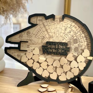 Peut inclure: Livre d'or en bois en forme de Faucon Millenium, avec un cadre et un support noirs. Le design comprend des détails gravés et le logo Star Wars avec les noms "Sherri & David" et la date "4 mai 2024". Petits coeurs en bois inclus.