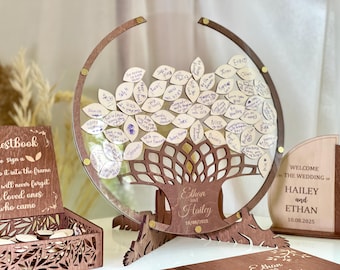 Puede incluir: Un libro de visitas de boda de madera con forma de árbol con etiquetas en forma de hoja para que los invitados firmen. El árbol es marrón con un marco circular y los nombres "Ethan y Hailey" y la fecha de la boda están grabados.