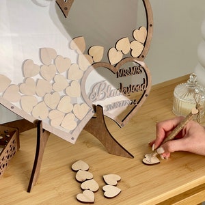 May include: A wooden heart-shaped wedding guest book box with a clear acrylic lid. The box is filled with small wooden hearts for guests to write messages on. The box is decorated with the words "Mr. & Mrs. Blackwood" and the date "30/07/2024".