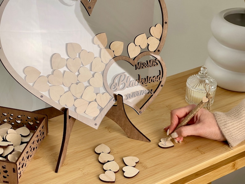 May include: A wooden heart-shaped wedding guest book box with a clear acrylic lid. The box is filled with small wooden hearts for guests to write messages on. The box is decorated with the words "Mr. & Mrs. Blackwood" and the date "30/07/2024".