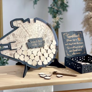 Könnte beinhalten: Eine hölzerne Star Wars-Themen-Gästebuch-Alternative. Das Design zeigt eine Millennium Falcon-Form mit Platz für Holzherzen. Enthält eine schwarze Box mit einem Schild mit der Aufschrift "Guestbook". Das Gästebuch steht auf einem Holztisch.