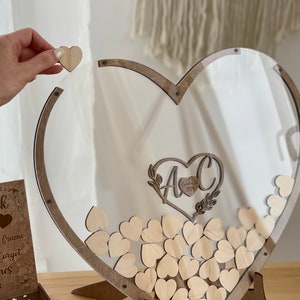 May include: A wooden heart-shaped frame with a clear acrylic insert. The frame is filled with small wooden hearts. The frame has a sign that says "Guestbook" and "Please sign a heart and drop it into the frame. We will never forget our loved ones who came."