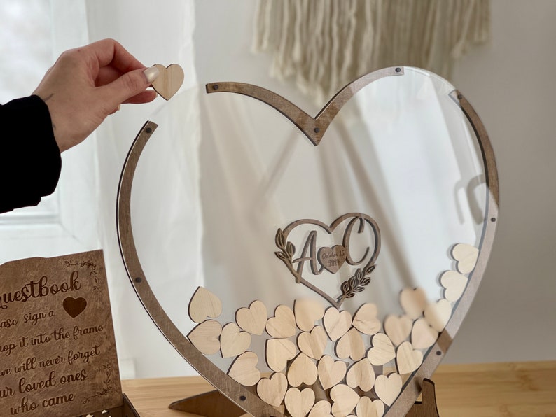 May include: A wooden heart-shaped frame with a clear acrylic insert. The frame is filled with small wooden hearts. The frame has a sign that says "Guestbook" and "Please sign a heart and drop it into the frame. We will never forget our loved ones who came."