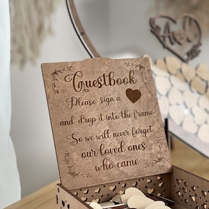 May include: A wooden guest book box with a sign that reads "Questbook Please sign a and drop it into the frame So we will never forget our loved ones who came". The box is decorated with a heart and floral designs.