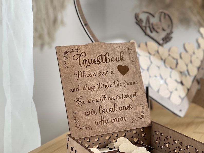 May include: A wooden guest book box with a sign that reads "Questbook Please sign a and drop it into the frame So we will never forget our loved ones who came". The box is decorated with a heart and floral designs.