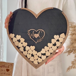 May include: A black heart-shaped wooden frame with a clear acrylic insert. The frame is decorated with a wooden heart with the initials "A & C" and the date "October 28, 2023". The frame is filled with small wooden hearts that guests can sign and drop into the frame.