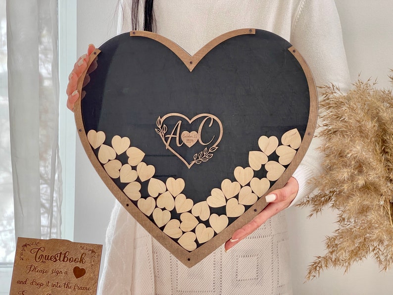 May include: A black heart-shaped wooden frame with a clear acrylic insert. The frame is decorated with a wooden heart with the initials "A & C" and the date "October 28, 2023". The frame is filled with small wooden hearts that guests can sign and drop into the frame.