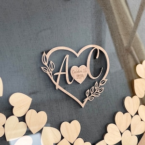 May include: A wooden heart-shaped guest book with the initials "A & C" and the date "October 15, 2024" engraved on it. The heart is surrounded by smaller wooden hearts.