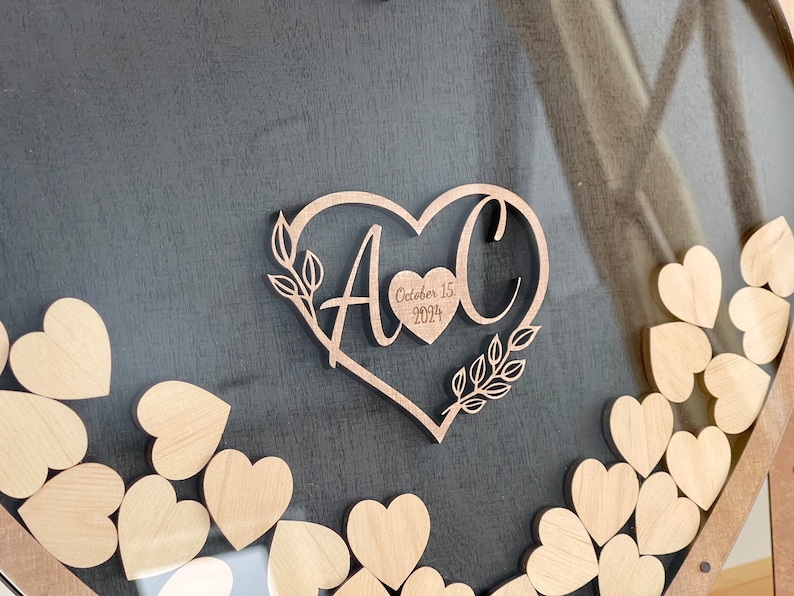 May include: A wooden heart-shaped guest book with the initials "A & C" and the date "October 15, 2024" engraved on it. The heart is surrounded by smaller wooden hearts.