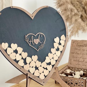 May include: A wooden heart-shaped guest book frame with a black chalkboard background. The frame has a cutout of a heart with the initials "A & C" and the date "October 12, 2023". The frame is filled with small wooden hearts that guests can write messages on and drop into the frame.