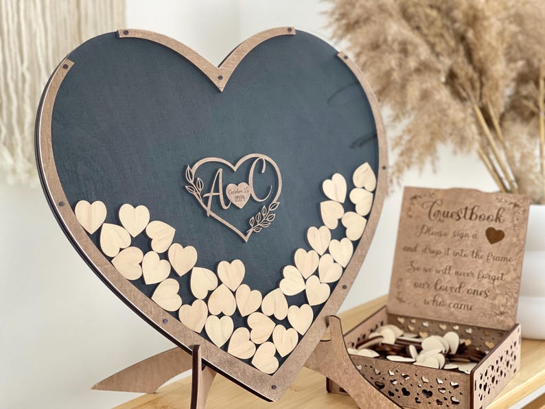 May include: A wooden heart-shaped guest book frame with a black chalkboard background. The frame has a cutout of a heart with the initials "A & C" and the date "October 12, 2023". The frame is filled with small wooden hearts that guests can write messages on and drop into the frame.