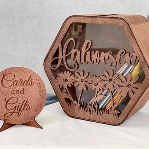 Könnte beinhalten: Holzkartenbox mit floralem Design und dem ausgeschnittenen Namen "Halvorsen". Ein passendes rundes Schild mit der Aufschrift "Karten und Geschenke" ist ebenfalls vorhanden. Die Box ist sechseckig mit einer transparenten Vorderseite.