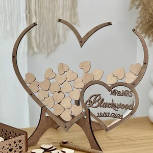 May include: Wooden heart-shaped wedding guest book with a clear acrylic insert. The hearts are decorated with the names "MR & MRS Blackwood" and the date "10/07/2024".