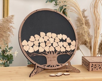 Puede incluir: Un libro de visitas de madera con forma de árbol con fondo negro. El árbol tiene ramas con corazones de madera que los invitados pueden firmar. El texto "Joe & Ashley" y "July 11, 2023" está grabado en el árbol.