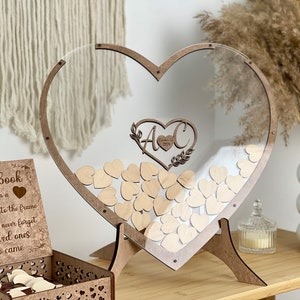 May include: A wooden heart-shaped frame with a clear acrylic insert. The frame is standing on a wooden stand. The frame is filled with small wooden hearts. The frame has a sign that says "Guestbook" and "Please sign a heart and drop it into the frame so we will never forget our loved ones who came".