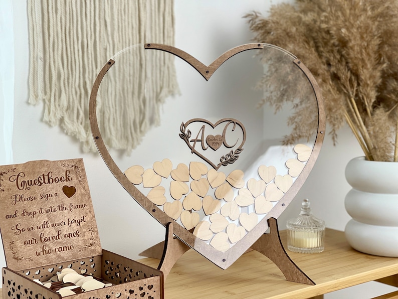 May include: A wooden heart-shaped frame with a clear acrylic insert. The frame is standing on a wooden stand. The frame is filled with small wooden hearts. The frame has a sign that says "Guestbook" and "Please sign a heart and drop it into the frame so we will never forget our loved ones who came".