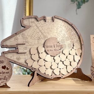 May include: A wooden wedding guest book shaped like the Millennium Falcon from Star Wars. The guest book has a slot for guests to drop wooden hearts into. The guest book is personalized with the names "Sherri & David" and the date "4th May, 2024".