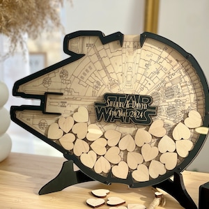May include: A black wooden Star Wars Millennium Falcon guest book with a cutout for wooden hearts. The guest book is personalised with the names "Sherri & David" and the date "4th May, 2024".