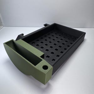 May include: A two-tone organizer with a black rectangular tray and an olive green curved compartment. The tray has a grid of square holes. The green section has a cylindrical hole and a rectangular slot. The organizer is designed for desk or workspace organization.