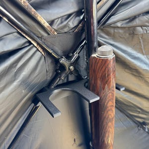 May include: A black metal gun holder with a brown wooden shotgun resting in it. The holder is attached to the inside of a camouflage hunting blind.