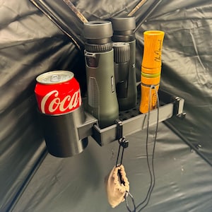May include: A black plastic holder with a can of Coca-Cola, two pairs of binoculars, and a wooden duck call. The holder is attached to a camouflage fabric.