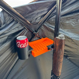May include: A black and orange hunting blind accessory with a cup holder, a shell holder, and a gun holder. A can of Coca-Cola is in the cup holder.