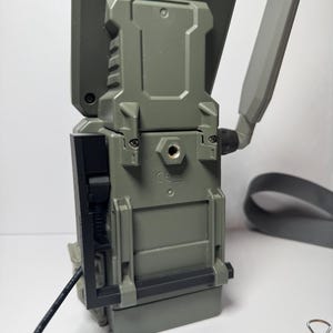 May include: A gray and black trail camera with a rectangular body and a hinged door on the side. It has a gray antenna and a gray strap. A black power cord is connected to the side.