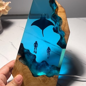 May include: A vertical rectangular lamp made of natural wood and translucent blue resin. The resin section features embedded silhouettes of a manta ray and two scuba divers. The wood has a visible grain pattern and irregular edges bordering the resin.