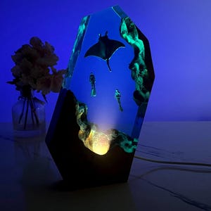 May include: A tall, angular resin lamp featuring an underwater scene with a manta ray and two scuba divers suspended in a translucent blue medium. The base incorporates dark wood elements and a glowing light source that illuminates the interior.