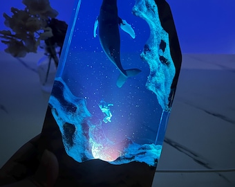 May include: A whale and a small sea creature are encased in a clear blue resin block with a dark wood base. The resin has a glittery effect and the whale is silhouetted against the blue background.
