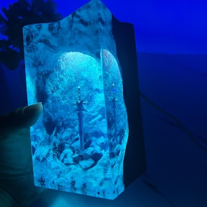 May include: A blue resin lamp with two swords embedded inside. The lamp is lit from within, casting a blue glow.