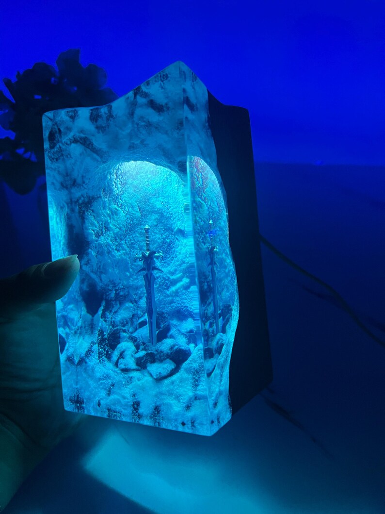 May include: A blue resin lamp with two swords embedded inside. The lamp is lit from within, casting a blue glow.