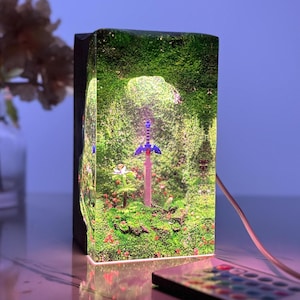 Puede incluir: Lámpara rectangular con una escena de bosque en miniatura y una espada. La lámpara tiene un marco negro y una parte frontal transparente, mostrando un diorama detallado con follaje verde, flores rojas y una espada brillante. Un mando a distancia está en primer plano.
