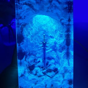 May include: A blue resin sculpture of a sword in a cave. The sword is silver and has a detailed design. The cave is made of clear resin and has a blue glow.