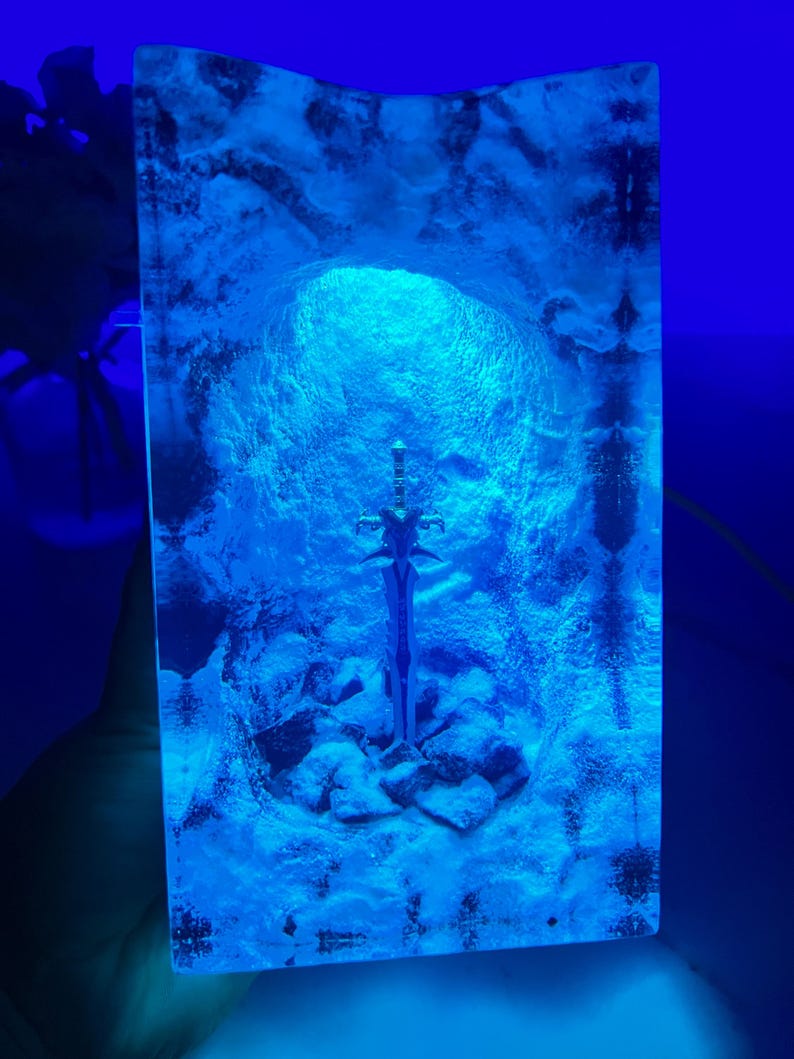 May include: A blue resin sculpture of a sword in a cave. The sword is silver and has a detailed design. The cave is made of clear resin and has a blue glow.