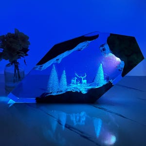 Puede incluir: Un pisapapeles de resina geométrica transparente con un diseño grabado en 3D de una escena de invierno con dos ciervos y pinos. El pisapapeles está iluminado desde atrás con una luz azul.