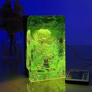 May include: A rectangular, illuminated resin lamp with a vibrant green interior, resembling a miniature landscape. The lamp has a black base and is controlled by a remote. A clear glass vase with flowers is in the background.
