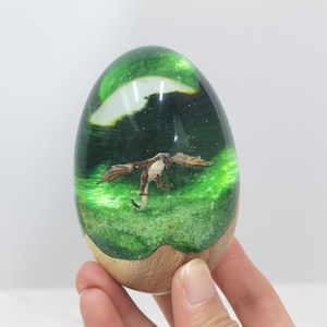 Puede incluir: Un huevo de resina verde con una pequeña figura de dragón en su interior. El huevo está sobre una base de madera.