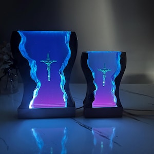 May include: Two resin lamps with a black base and a clear resin body. The resin body is filled with a blue and purple liquid and a small silver cross with a figure of Jesus Christ. The lamps are illuminated from within, creating a soft glow.