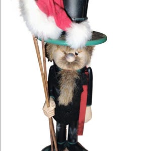 May include: A whimsical chimney sweep figurine with a black top hat, green brim, and a red scarf. The figurine holds wooden rods and a red mitten with white trim. The figure has a furry beard and a neutral expression.